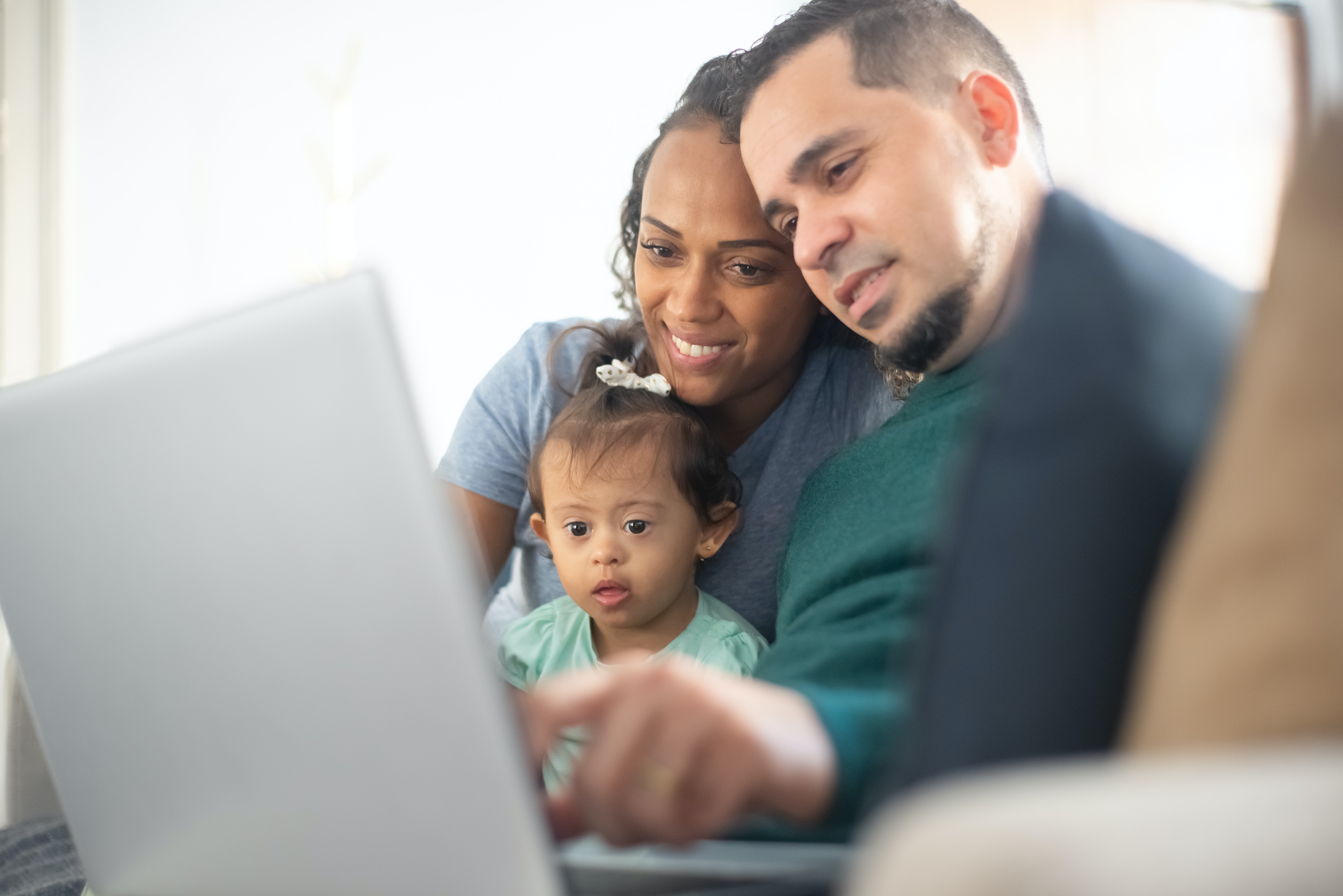 A family is pictured learning together on a laptop. A Black mother in her 30’s holds her daughter in her lap. Her young daughter, who has Down Syndrome, looks to be around the age of two and she is looking closely at the computer. A Latino father in his 30’s is pointing to the screen to show his wife and daughter something.